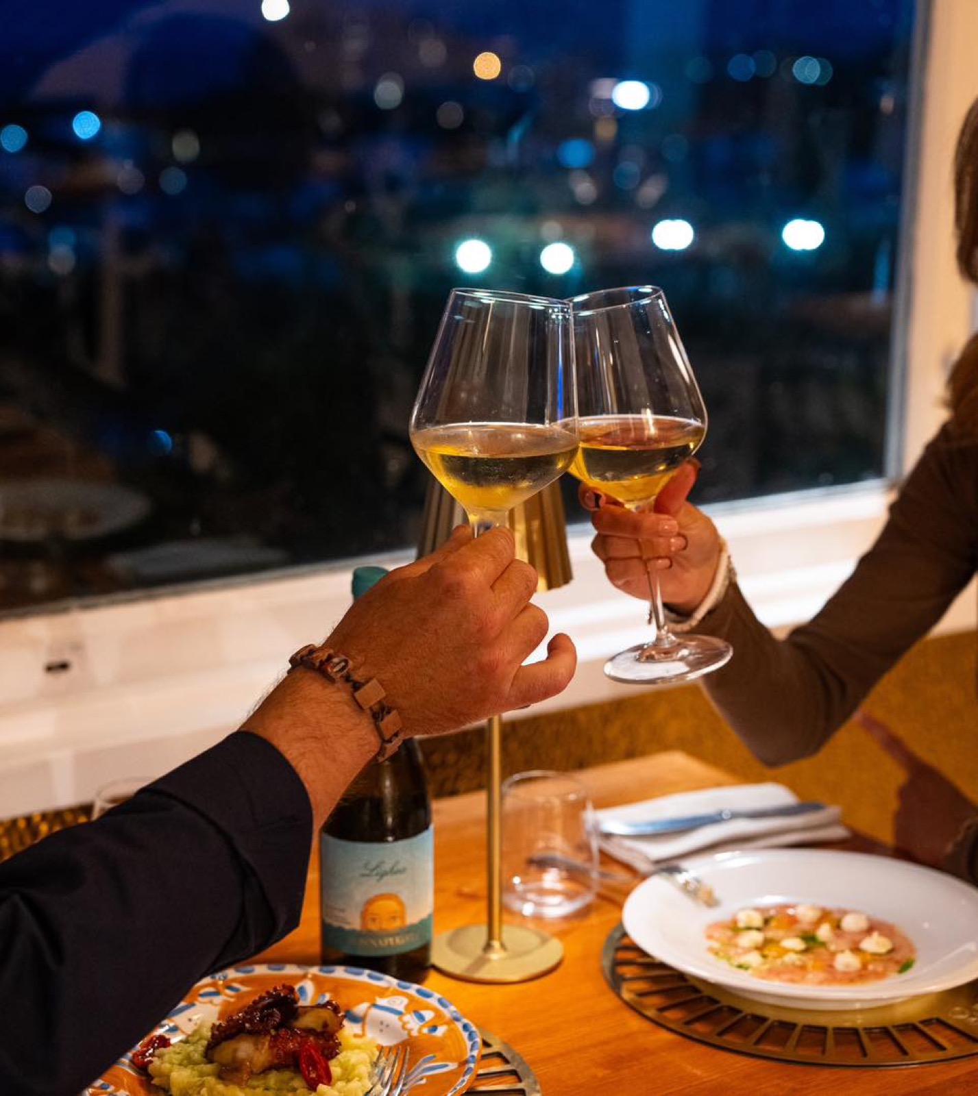 Brindisi con calici di vino bianco al tavolo, con le luci della sera sul golfo